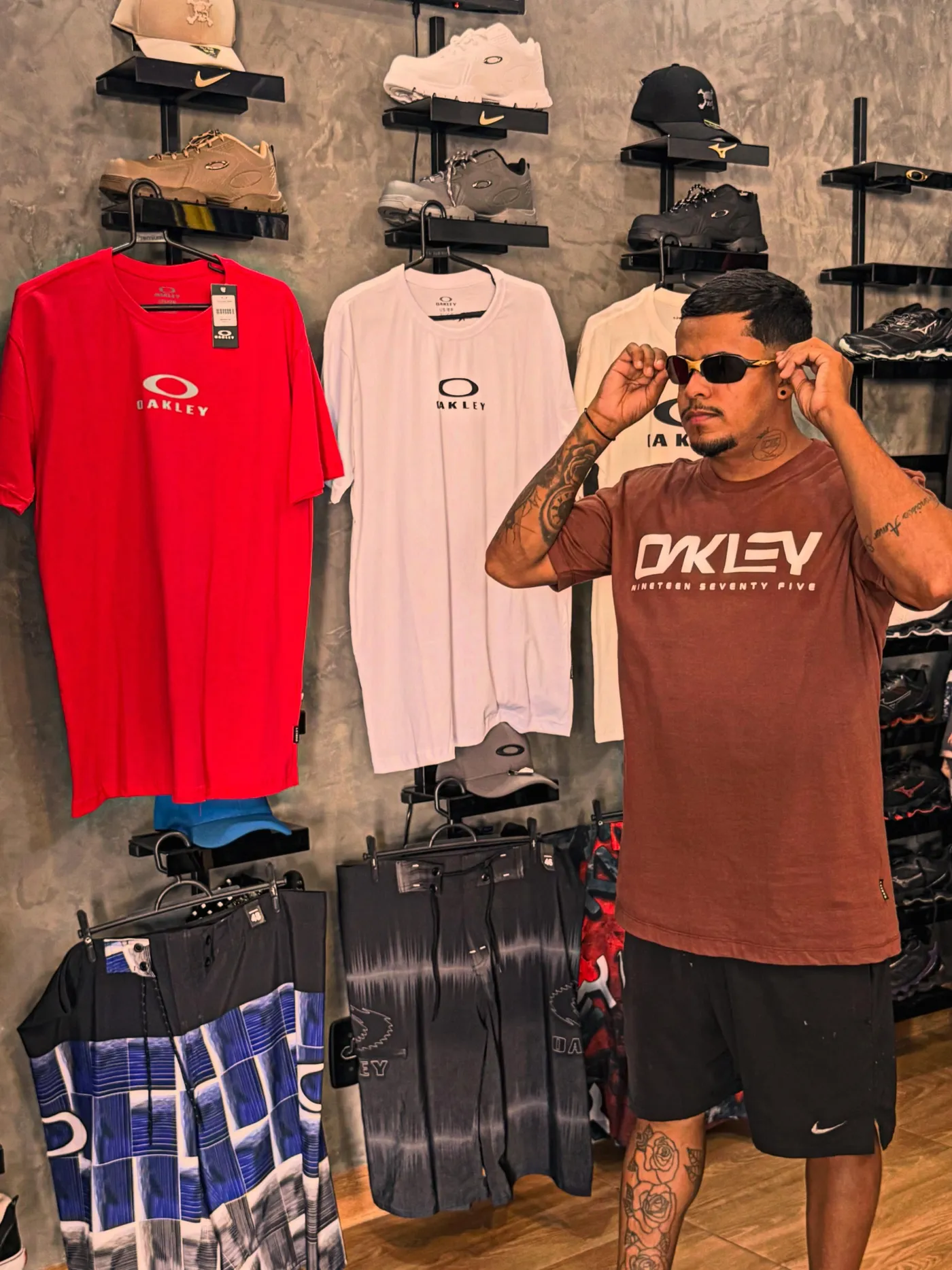 Na DK Modas, um rapaz de camiseta Oakley marrom ajeita os óculos na frente da parede de cimento, com camisetas e bermudas Oakley penduradas e tênis nas prateleiras pretas